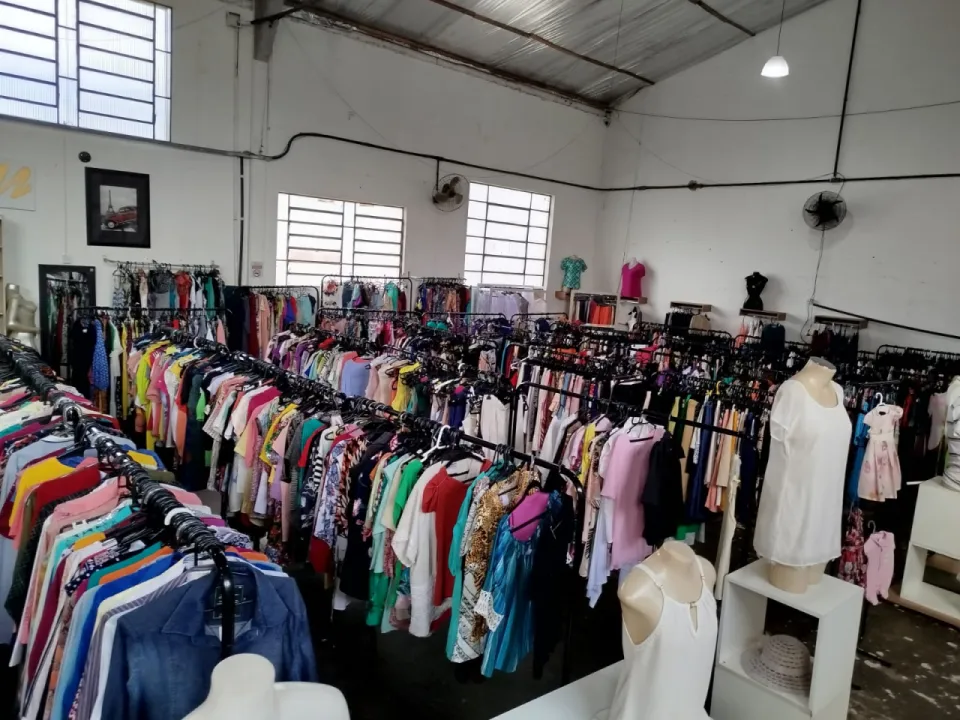 Vista geral do salão do bazar, com fileiras de araras de roupas, manequins e janelas altas